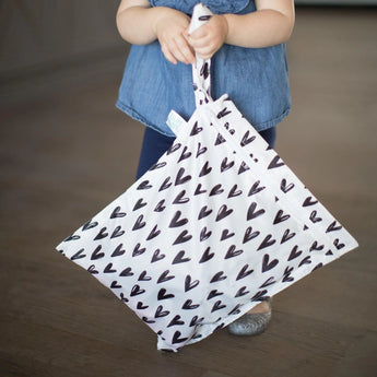 A child holds Bumkins Wet Bag: Hearts, a folded umbrella with black hearts on waterproof fabric against a white background.