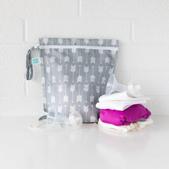 Bumkins Wet Bag: Arrow in waterproof fabric next to cloth diapers and nursing gear on a white surface against a white brick wall.