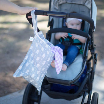 A baby sits in a stroller outdoors as a hand holds Bumkins Wet Bag: Arrow, featuring a purple cloth attached to the handle.