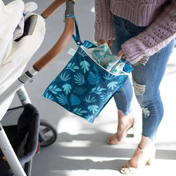 A person places items into the Bumkins Wet Bag: Blue Tropic attached to a stroller, wearing jeans and a purple sweater. The fabric shines.