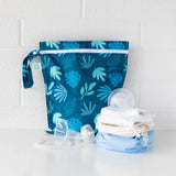 The Blue Tropic Wet Bag, baby cloth diapers, and Bumkins SuperBib sit on a white surface with a brick backdrop.