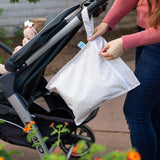 A person attaches a Bumkins Wet Bag: Wander to a stroller with a baby outdoors. Orange flowers bloom nearby.