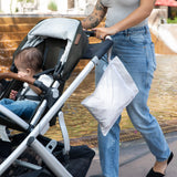 A tattooed person in jeans and a gray top pushes a stroller by the fountain, carrying a Bumkins Wet Bag: Wander with a toddler.