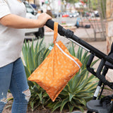 Person pushing a stroller with the Bumkins Wet Bag: Grounded, featuring an orange pattern, plants and street scene in the background.