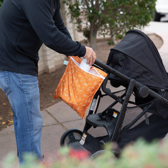 Someone attaches a Bumkins Wet Bag: Grounded (orange pattern) to a black stroller on a paved path outside.