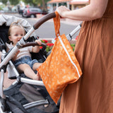 A woman in a brown dress pushes a stroller with a baby and carries a Bumkins Wet Bag: Grounded.