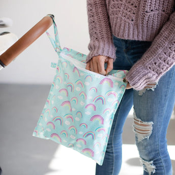 A person in a pink sweater and ripped jeans hangs a Bumkins Wet Bag: Rainbows on a stroller handle.