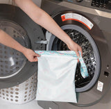 Someone places a Bumkins Rainbows Wet Bag, made of waterproof fabric, into a front-load washer with a laundry basket nearby.