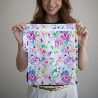 Person holding a Bumkins Watercolor wet bag with a floral pattern in pastel pink and purple, made of waterproof fabric.