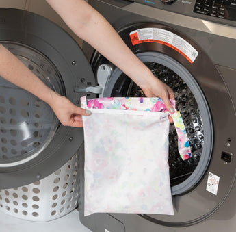 A person loads a Wet Bag: Watercolor by Bumkins, made of waterproof fabric, into a front-loading washing machine.