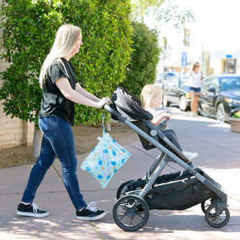 A woman pushes a child in a stroller on a sunny sidewalk, with Bumkins Wet Bag: Ocean Life slung over her shoulder.