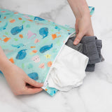 Hands placing clothes into Bumkins Wet Bag: Ocean Life, a waterproof bag with cartoon sea creatures, on a white surface.