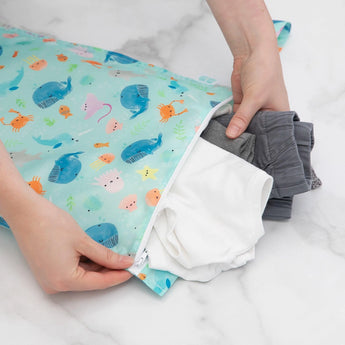 Hands placing clothes into Bumkins Wet Bag: Ocean Life, a waterproof bag with cartoon sea creatures, on a white surface.