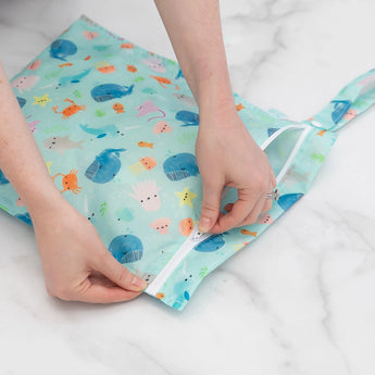 Hands zip a Bumkins Wet Bag: Ocean Life in light blue, featuring colorful sea creatures like whales, starfish, and crabs on waterproof fabric. The background is white.