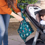 A woman expertly attaches a Bumkins Wet Bag: Jungle to a stroller as she strolls outside with her child.