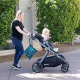 A woman pushes a stroller with her Bumkins Wet Bag: Jungle neatly packed; trees and a building are in the background.