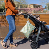 Woman in orange shirt pushes child in stroller near a fountain, carrying Bumkins Wet Bag: Camp Gear under bright sun.