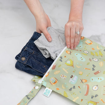 Hands placing gray shirt and denim jeans into Bumkins Wet Bag: Camp Gear with a green camping-themed pattern on a white surface.