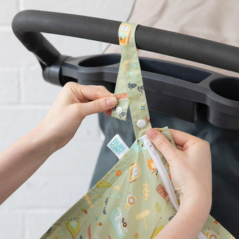 Hands attach the Bumkins Wet Bag: Camp Gear, a green patterned waterproof cloth with snap button straps, to a stroller.