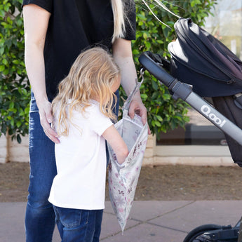 A child peeks into a Bumkins Wet Bag: Floral, made of waterproof fabric, held by an adult next to a stroller outdoors.