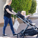 A woman walks with a toddler in a stroller, featuring Bumkins Wet Bag: Floral, with waterproof fabric, on a sidewalk near greenery.