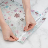 Hands closing a Bumkins Wet Bag: Floral on marble, made of waterproof fabric with a zipper.