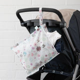 A Bumkins Wet Bag: Floral, made of waterproof fabric, hangs on a stroller, holding a white shirt and jeans.