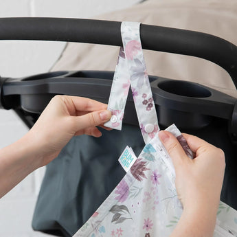 Hands attaching the Bumkins Wet Bag: Floral to a stroller with snap buttons, crafted from waterproof fabric.