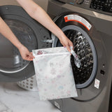 Person places a Bumkins Wet Bag: Floral into a front-loading washer, with a waterproof fabric laundry basket nearby.