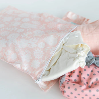 The Bumkins Lace Wet Bag in pink floral with a white zipper holds two pastel polka dot reusable diapers, made from waterproof fabric.