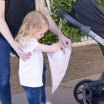 A child puts an item in a Bumkins Wet Bag: Lace attached to a stroller. The pink bag is reusable and waterproof.