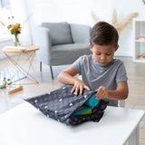 A child in a gray shirt opens a Bumkins Mickey Mouse Icon Black + White wet bag at a table in a cozy living room with flowers.