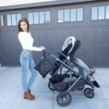 A person in a white sweater pushes a double stroller by a garage, holding a Bumkins Wet Bag: Mickey Mouse Icon Black + White.