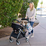 A smiling woman in jeans pushes a stroller past greenery, holding a Bumkins Wet Bag: Mickey Mouse Classic.