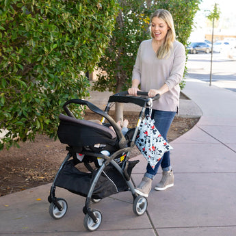 A smiling woman in jeans pushes a stroller past greenery, holding a Bumkins Wet Bag: Mickey Mouse Classic.