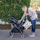 A woman with long hair leans over a black and gray baby stroller on the sidewalk, smiling while wearing a light sweater, jeans, and gray shoes. A Bumkins Wet Bag: Mickey Mouse Classic hangs from the stroller handle. Green foliage in the background creates a serene setting.