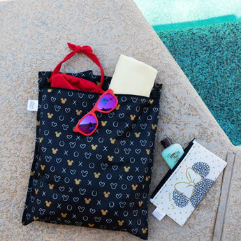 A black patterned bag with red sunglasses, a yellow towel, and a red headband lounges by the shimmering pool. Nearby, Bumkins Wet Bag: Minnie Mouse Icon Black + White, crafted from waterproof fabric with a bow design, sits beside a bottle marked ocean-bound plastic.