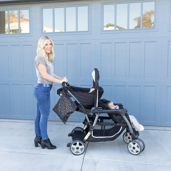 A person smiles with a child in a stroller by a blue garage, holding Bumkins colorful Minnie Mouse Icon Black + White wet bag.