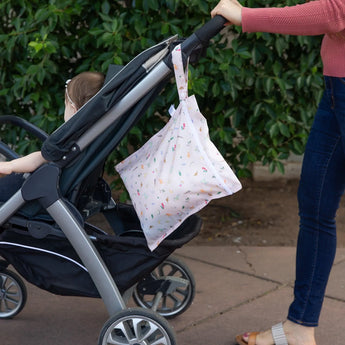 A person strolls with a baby stroller and Bumkins Wet Bag: Princess Magic, along a sidewalk lined with greenery.