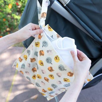 Hands place a cloth into a Bumkins Winnie and Friends wet bag, hanging from a stroller handle outdoors.