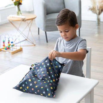 A boy in a gray shirt sits at a white table, unwrapping a Bumkins Super Mario™ Lineup wet bag in a modern room with toys.