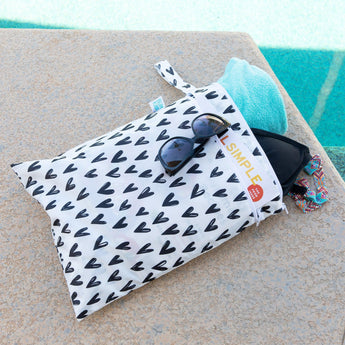 The Bumkins Wet/Dry Bag: Hearts, made with waterproof fabric, holds sunglasses, a turquoise towel, and colorful jewelry by the pool.