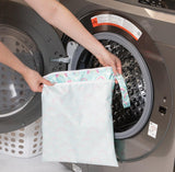 A person places a Bumkins Wet/Dry Bag: Rainbows into an open washer, with a laundry basket beside it.