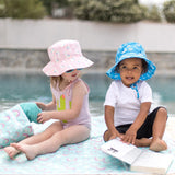 Two kids in sun hats sit by the pool on a mat. One reads, while the other reaches into a Bumkins Wet/Dry Bag: Rainbows.