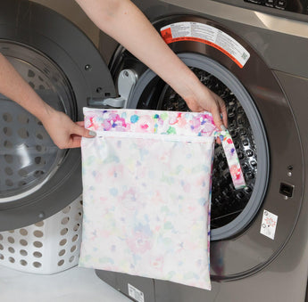 Someone places the Bumkins Wet/Dry Bag: Watercolor into a front-loading washer, with a laundry basket nearby.