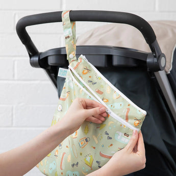 A hand attaches a Bumkins Wet/Dry Bag: Camp Gear with camping prints to a stroller, ideal for travel or waterproof needs.