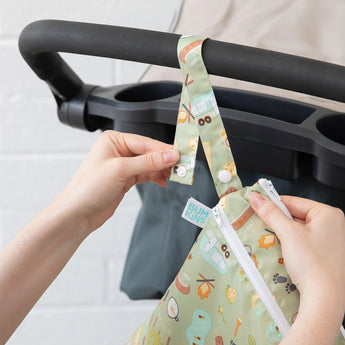 A person attaching a Bumkins Wet/Dry Bag: Camp Gear, crafted from waterproof fabric, to a stroller handle using a snap button strap.