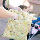 A hand holds the Bumkins Wet/Dry Bag: Camp Gear near a child in a stroller outdoors.