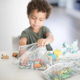 A curly-haired child opens a Bumkins Clear Travel Bag with cacti design, revealing toys at a table filled with more toys and blocks.