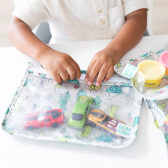 A child plays with toy cars and playdough on a white table near a Bumkins Clear Travel Bag in the cactus design.